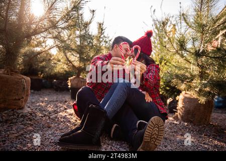 Couple amoureux en chemises à carreaux, chapeaux tricotés tiennent des cannes de bonbons en forme de cœur dans les mains près du marché vert des arbres de Noël. L'homme et la femme s'embrassent, rient, h. Banque D'Images