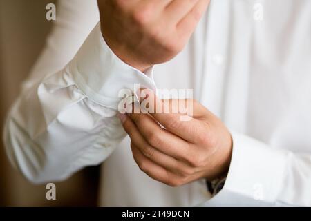 Gros plan des mains fixant un bouton de manchette argenté sur une chemise blanche. Élégance et préparation sous lumière naturelle. Banque D'Images