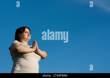 Portrait de femme d'âge moyen contre ciel bleu. Femme calme tenant les mains dans le mudra namaste pendant la méditation. Femme en surpoids pratique le yoga à l'extérieur Banque D'Images