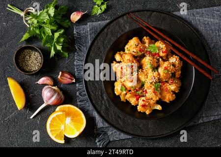 bouquets de chou-fleur d'orange croustillants saupoudrés de graines de sésame, de flocons de piment et de persil dans un bol noir sur une table en béton avec des ingrédients et une baguette Banque D'Images