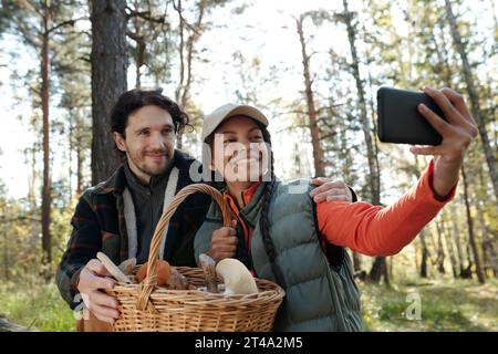 Jeunes dates interculturelles avec panier de champignons regardant la caméra smartphone avec des sourires pendant selfie dans la forêt d'automne Banque D'Images