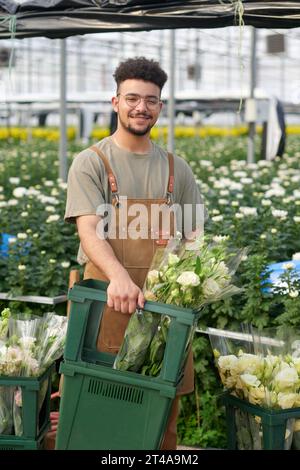 Heureux jeune homme travailleur de serre industrielle avec des grappes de fleurs fraîches d'eustoma dans la boîte debout devant la caméra et vous regardant Banque D'Images