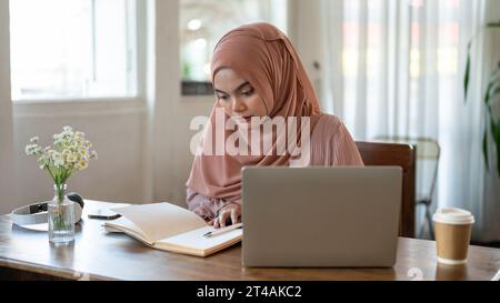 Une belle et concentrée jeune femme asiatique-musulmane lit un livre tout en travaillant à distance dans un café le week-end. Banque D'Images