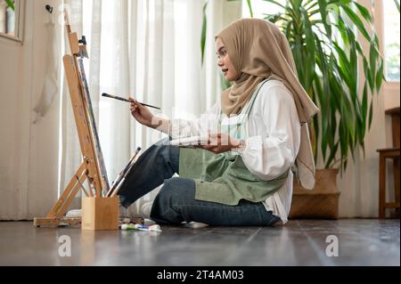 Une image de côté d'une artiste féminine asiatique-musulmane créative et concentrée assise sur le sol dans son atelier et peint sur toile sur un chevalet. Banque D'Images