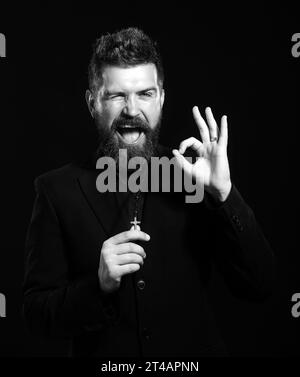 Jeune homme portant l'uniforme de prêtre debout sur fond blanc faisant geste ok avec la main souriante, oeil regardant à travers les doigts avec le visage heureux. Banque D'Images