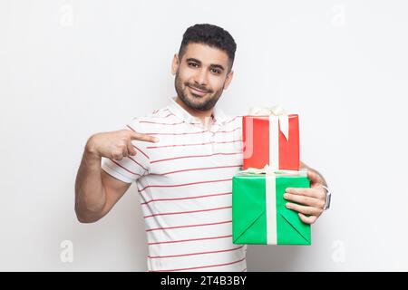 Portrait d'un beau jeune homme barbu positif et joyeux portant un t-shirt rayé debout regardant la caméra, pointant vers deux boîtes cadeaux. Studio intérieur tourné isolé sur fond gris. Banque D'Images