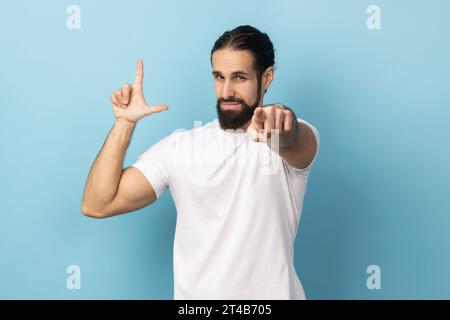 Portrait d'un homme frustré avec barbe portant un T-shirt blanc montrant un geste plus lâche et pointant vers la caméra, regardant avec un visage grincheux. Studio intérieur tourné isolé sur fond bleu. Banque D'Images