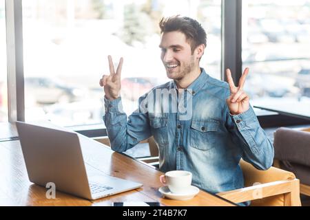 Portrait de souriant heureux homme freelancer dans la chemise bleu jeans travaillant sur ordinateur portable, a appel vidéo, montrant v signe à l'écroche, se réjouit de sa victoire. Prise de vue intérieure près de la grande fenêtre, fond de café. Banque D'Images