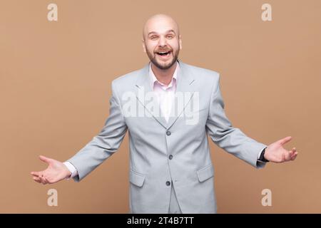 Portrait de l'homme barbu chauve attrayant rit joyeusement à l'histoire drôle ou à la blague, être de bonne humeur, exprimer des émotions positives, porter une veste grise. Studio intérieur tourné isolé sur fond brun. Banque D'Images