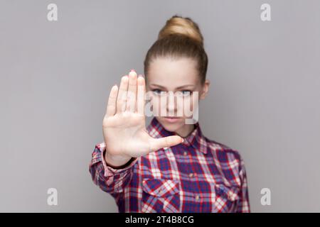 Portrait de femme autoritaire sérieuse avec coiffure de chignon montrant le geste d'interdiction, montre le signe de paume d'arrêt, regardant la caméra, portant la chemise à carreaux. Studio intérieur tourné isolé sur fond gris. Banque D'Images