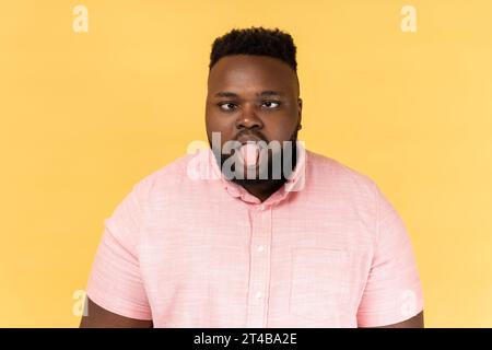 Portrait d'homme drôle fou désobéissant portant une chemise rose montrant la langue dehors et croisant ses yeux mécontent, se comportant indisciplinairement méchant. Studio intérieur tourné isolé sur fond jaune. Banque D'Images