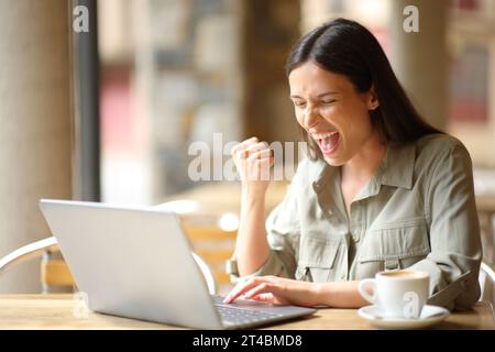 Femme excitée célébrant de bonnes nouvelles dans un restaurant vérifiant l'ordinateur portable Banque D'Images