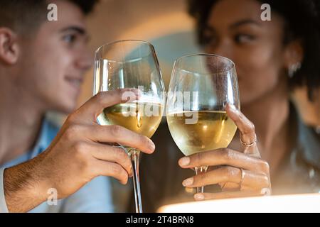 Un gros plan intime d'un jeune homme et d'une jeune femme grillant avec des verres de vin blanc. Leurs expressions ciblées et le cliquetis des lunettes symbolisent celebr Banque D'Images