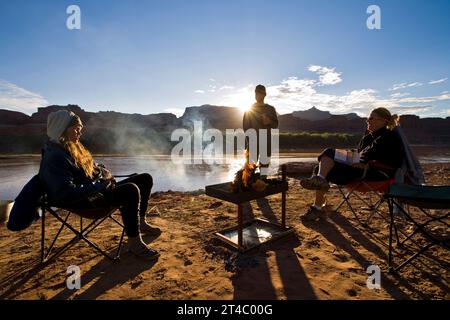 Trois personnes se sont rassemblées autour d'un feu au lever du soleil sur une plage de sable du fleuve Colorado dans l'Utah. Banque D'Images