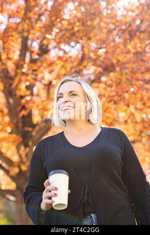 Femme aux cheveux blonds souriant, tenant le café au milieu du feuillage d'automne Banque D'Images