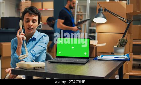 Femme entrepreneur répondant au téléphone fixe dans l'espace de magasin et en utilisant l'écran vert sur l'ordinateur portable. Femme discutant au téléphone avec cordon et regardant l'écran de maquette isolé. Banque D'Images