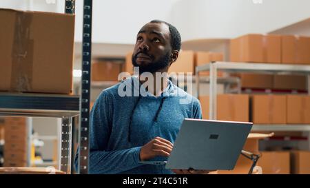 Homme afro-américain travaillant sur l'inventaire des marchandises en utilisant un ordinateur portable et prenant des notes dans l'entrepôt. Entrepreneur vérifiant les produits dans des boîtes en carton, travaillant sur le contrôle de la qualité et la distribution. Banque D'Images