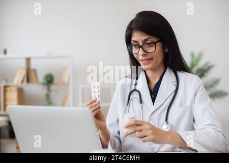 Travailleur de l'hôpital hindou parlant au patient sur appel vidéo et faisant la consultation sur le traitement dans le bureau lumineux. Infirmière attentive tenant des pilules en main Banque D'Images