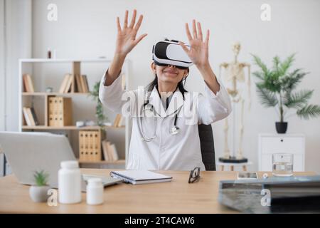 Infirmière indienne portant une veste de laboratoire en utilisant des lunettes de VR tout en étant assis au bureau avec des dispositifs modernes sur fond flou du cabinet. Thérapeute féminin préparant Banque D'Images