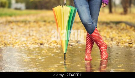 Une vue des jambes d'une femme dans des bottes en caoutchouc debout dans une flaque d'eau, appuyée sur un parapluie. Une femme debout dans une flaque d'eau entourée de feuilles d'automne tombées Banque D'Images