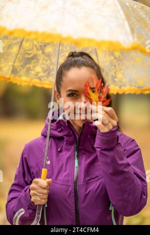 Jolie jeune femme sous un parapluie tenant une feuille d'érable colorée et couvrant son œil dans un parc d'automne. Banque D'Images
