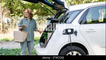 Femme avec sac à provisions à côté d'une voiture électrique de charge dans la cour d'une maison de campagne. Banque D'Images