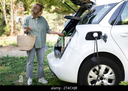 Femme avec sac à provisions à côté d'une voiture électrique de charge dans la cour d'une maison de campagne. Banque D'Images