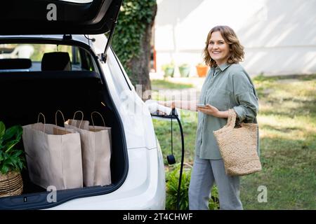 Femme avec sac à provisions à côté d'une voiture électrique de charge dans la cour d'une maison de campagne. Banque D'Images