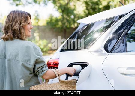 Femme avec sac à provisions à côté d'une voiture électrique de charge dans la cour d'une maison de campagne. Banque D'Images