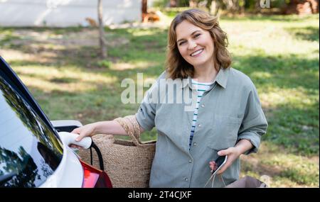 Femme avec sac à provisions à côté d'une voiture électrique de charge dans la cour d'une maison de campagne. Banque D'Images