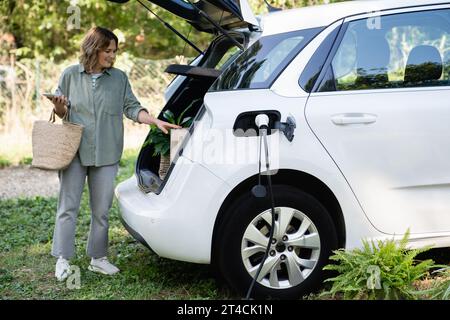 Femme avec sac à provisions à côté d'une voiture électrique de charge dans la cour d'une maison de campagne. Banque D'Images