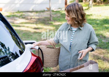 Femme avec sac à provisions à côté d'une voiture électrique de charge dans la cour d'une maison de campagne. Banque D'Images