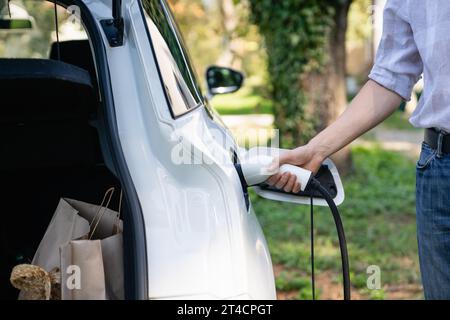 Un homme se tient près d'une voiture électrique et tient une prise de charge. Banque D'Images