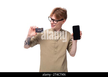 jeune étudiant européen inspiré aux cheveux rouges dorés habillé d'une chemise marron clair tenant une carte en plastique et un smartphone Banque D'Images