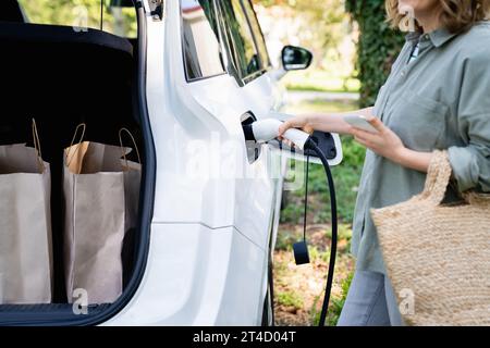 Femme avec sac à provisions à côté d'une voiture électrique de charge dans la cour d'une maison de campagne. Banque D'Images