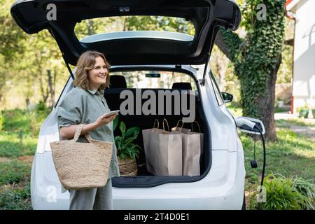 Femme avec sac à provisions à côté d'une voiture électrique de charge dans la cour d'une maison de campagne. Banque D'Images