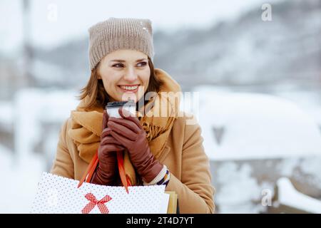 heureuse élégante femme d'âge moyen dans le chapeau brun et écharpe en manteau de chameau avec des gants, sacs à provisions et tasse de latte de soja dehors dans la ville en hiver. Banque D'Images