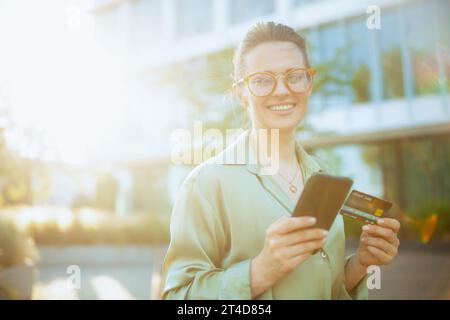 heureuse femme moderne de 40 ans employée dans le quartier des affaires en blouse verte et lunettes avec smartphone et carte de crédit. Banque D'Images