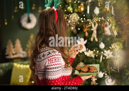 Noël. Vu de derrière élégante femme au foyer avec biscuit de Noël dans le pull de Noël traditionnel près de l'arbre de Noël à la maison moderne. Banque D'Images