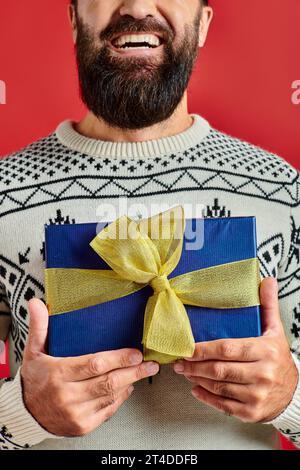 Homme barbu heureux coupé en pull d'hiver avec ornement tenant le cadeau de Noël sur fond rouge Banque D'Images