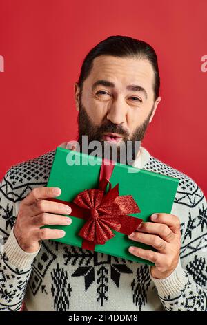 Air baiser de l'homme barbu en pull d'hiver avec ornement tenant le cadeau de Noël sur fond rouge Banque D'Images