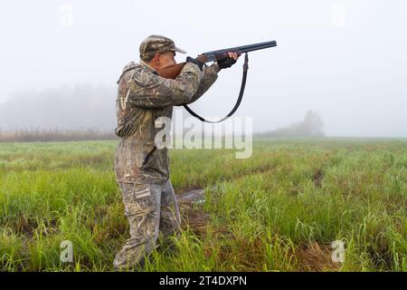 un homme vêtu de camouflage vise avec un fusil de chasse dans une prairie un matin brumeux Banque D'Images