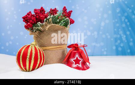 Houx baie rouge en toile de jute avec GUI, boule rouge et cloche sur la neige sur fond bleu avec des flocons de neige. Noël, nouvel an. Espace de copie Banque D'Images