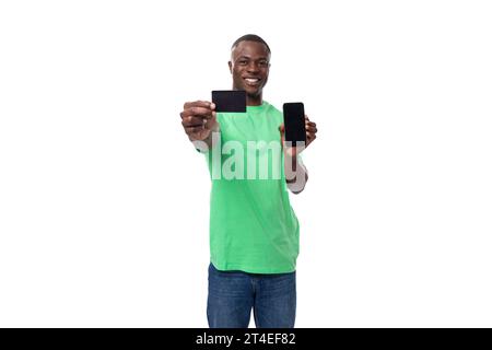 jeune homme africain bien entretenu dans un t-shirt vert clair et un jean montre une carte de crédit et un smartphone avec une maquette Banque D'Images
