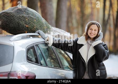 Happy Woman est en train d'écouter de la musique dans des écouteurs, de danser tout en emballant l'arbre de Noël avec le filet et les guirlandes sur un toit de sa voiture, se préparer à un Banque D'Images