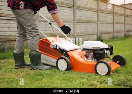 Un jardinier méconnaissable en gants de protection démarre le robot de tonte avant de couper de l'herbe verte dans son jardin. Homme avec tondeuse à gazon motorisé env Banque D'Images