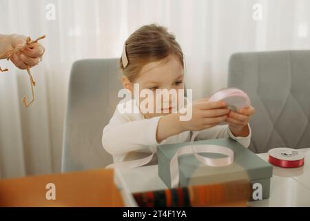 Belle petite fille prépare des cadeaux de Noël pour des amis et les emballe dans des boîtes tout en étant assis à la table Banque D'Images