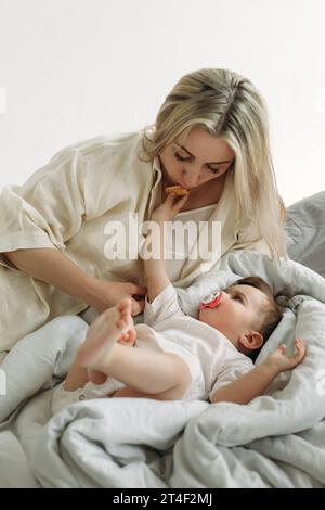 Une petite fille se couche dans son lit, suce une sucette, nourrit sa mère biscuits. Cadre vertical. Banque D'Images