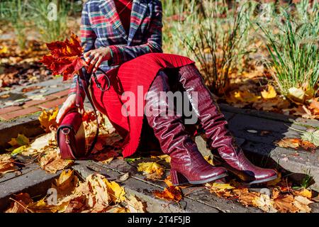 Gros plan de bottes hautes en cuir rouge bordeaux. Femme à la mode portant un blazer à carreaux élégant, robe assise dans le parc d'automne avec sac à main parmi l'automne Banque D'Images