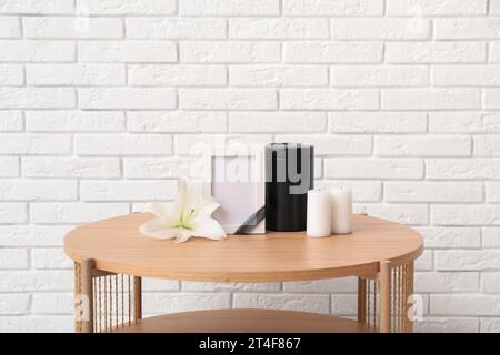 Cadre funéraire vierge, urne mortuaire, bougies et fleur de lys sur une table en bois près d'un mur de briques claires Banque D'Images